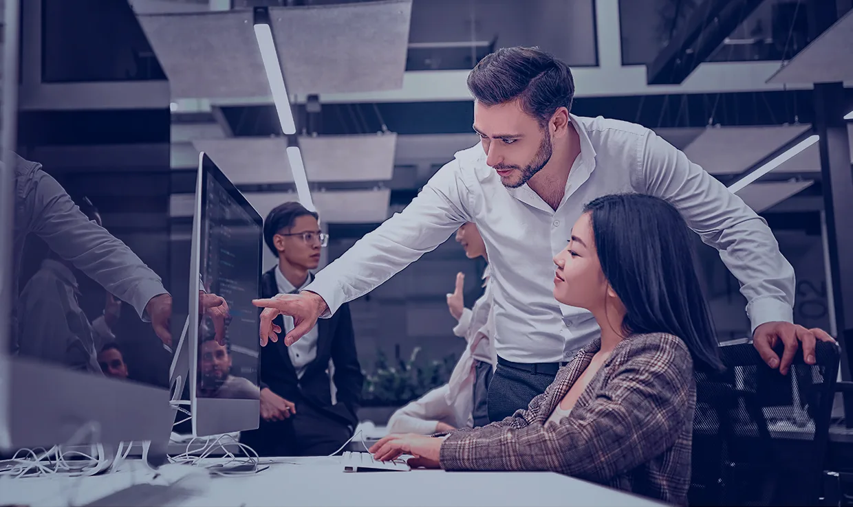 Man points at screen in an office. Female coworker watches. Professionals talk in workplace.