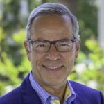 Close-up of a smiling man wearing glasses, a blue blazer, and a blue checkered shirt outdoors.