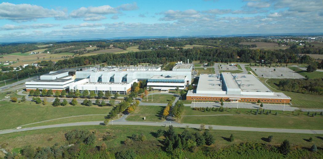 Aerial view of a modern university campus with white buildings, brick annex, and surrounding green fields and trees under a blue sky.