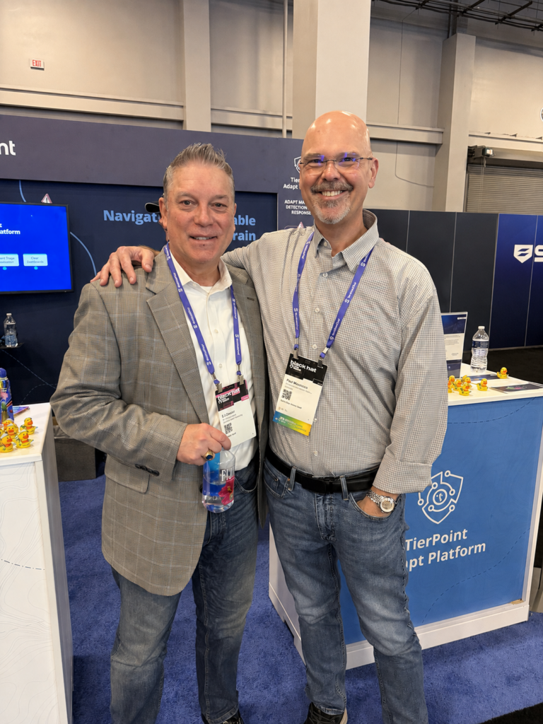 Two men at a tech conference pose with arms around each other, wearing lanyards and badges in front of a TierPoint booth.