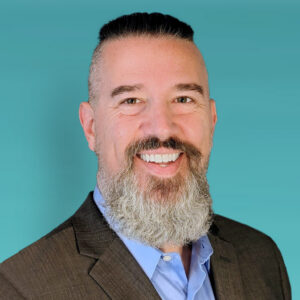 Smiling man with a gray beard wearing a brown suit and light blue shirt against a teal background; professional headshot.