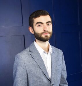 Portrait of a man with a beard in a light gray blazer and white shirt, posed against a dark blue paneled wall.
